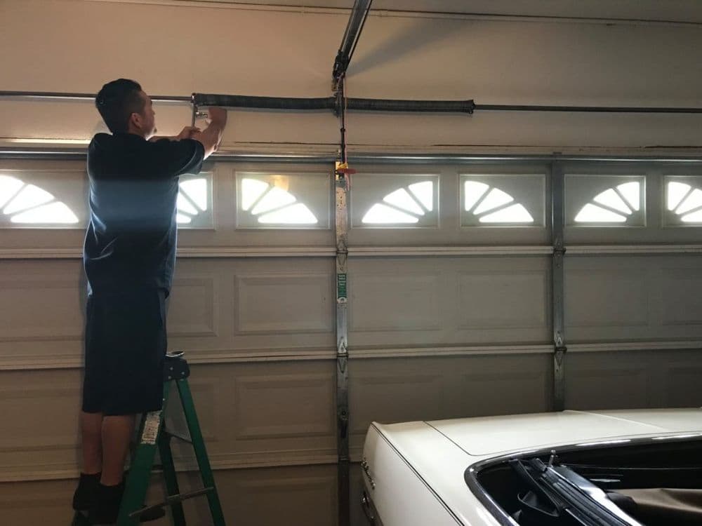 Man repairing a garage door spring while standing on a ladder next to a classic car.