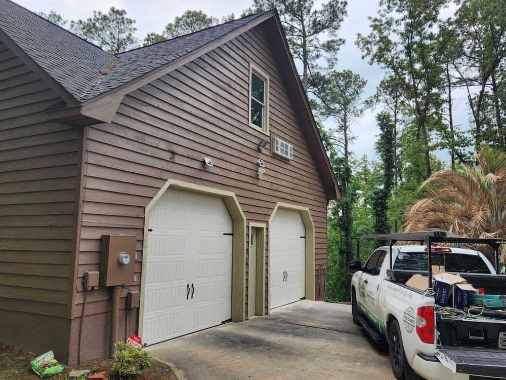 Two-car garage with wooden siding and a truck parked nearby, surrounded by greenery.