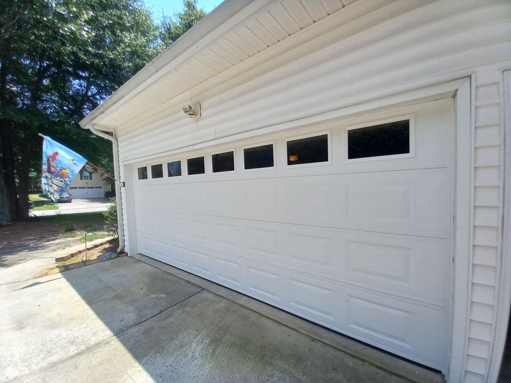 White garage door with decorative windows beside a driveway and a colorful flag.