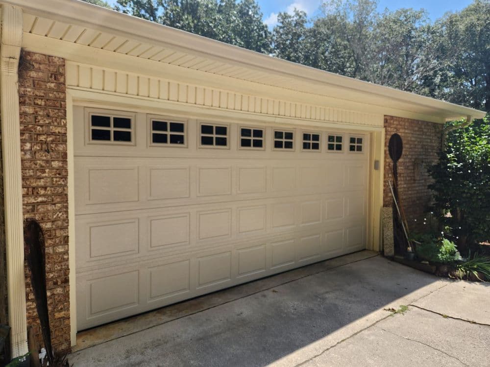 Modern beige garage door on a brick house with greenery and a stone driveway.