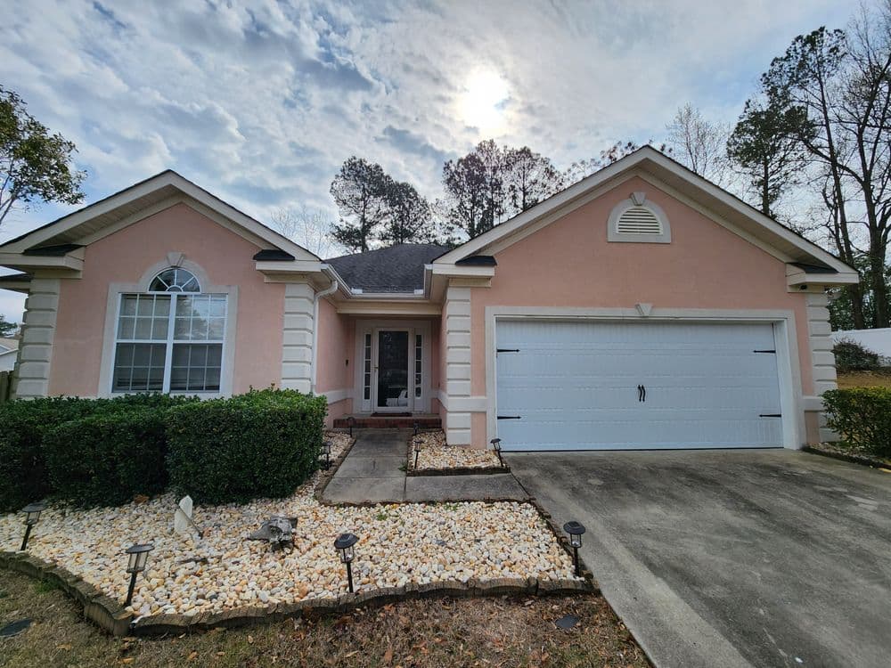 Pink single-story house with a garage, landscaped yard, and cloudy sky in the background.