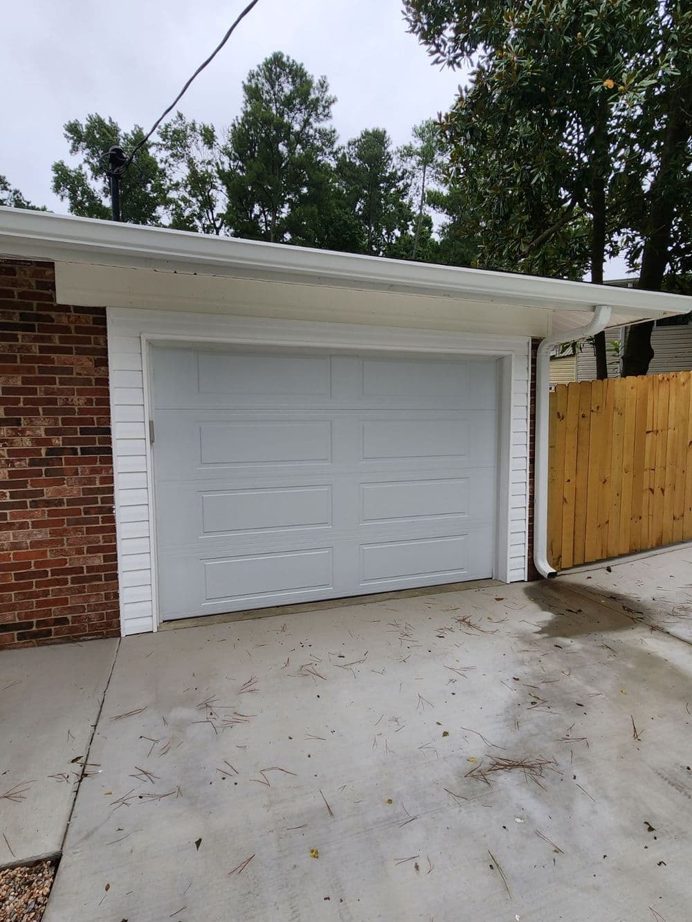 White garage door with raised panels beside a wooden fence amidst green trees.
