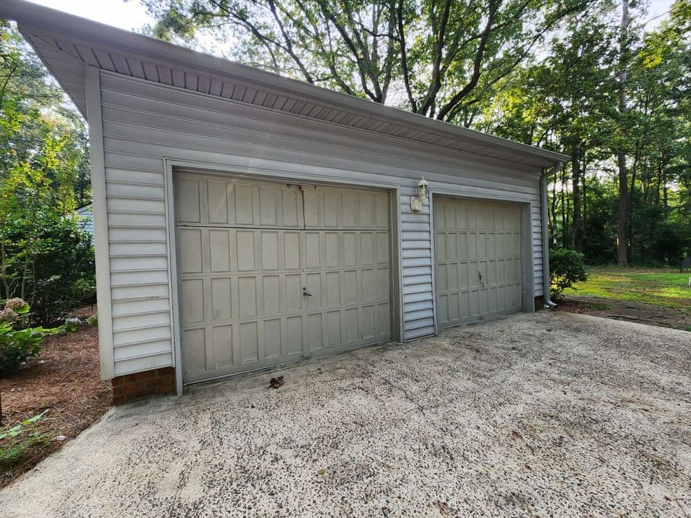 Two-car garage with a gray exterior and concrete driveway surrounded by trees.